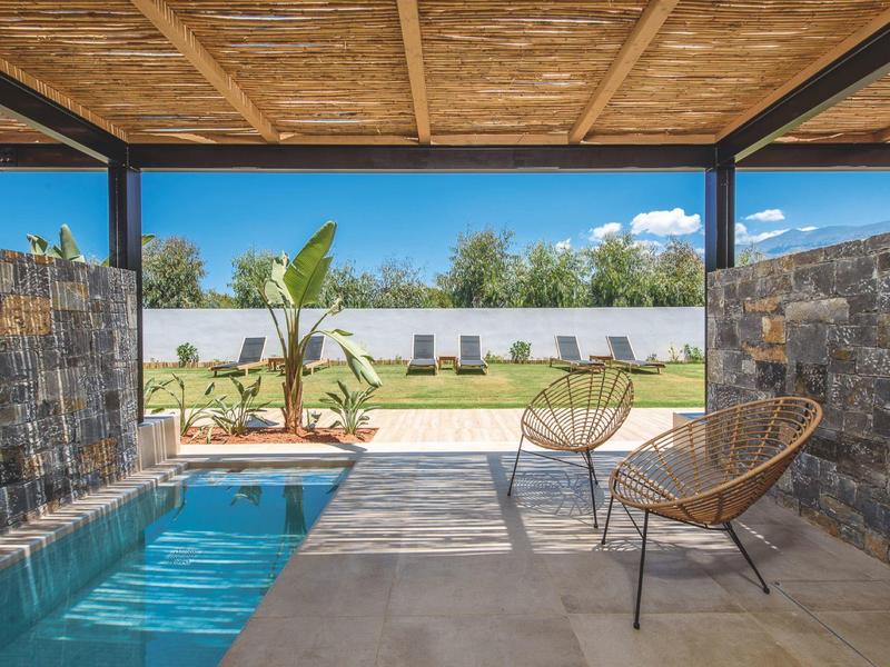 Overdekt zwembad met twee stoelen, waterval en ligstoelen buiten onder een blauwe lucht.