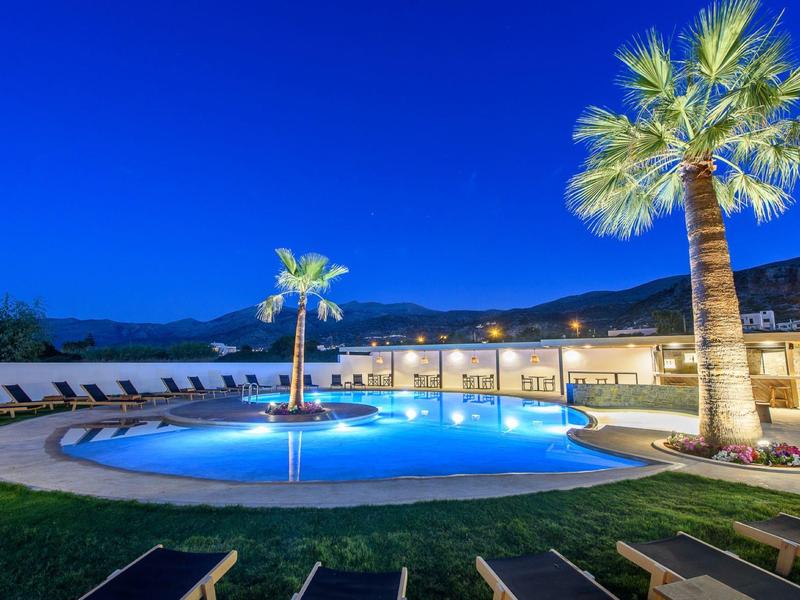 Verlicht zwembad met palmbomen en ligstoelen voor een hotel onder een heldere nachtelijke hemel.