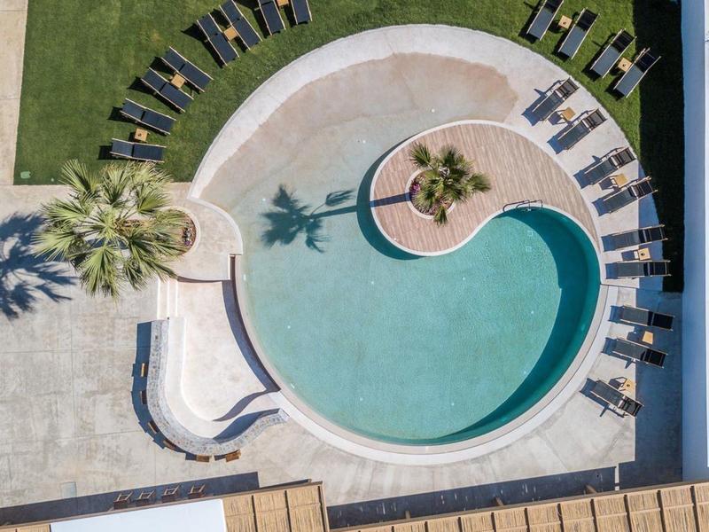 Rond zwembad met ligstoelen op gras en stenen tegels, palmbomen werpen schaduwen.