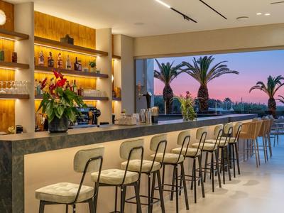 Bar d'hôtel moderne avec comptoir long, tabourets et vue sur des palmiers au coucher du soleil.