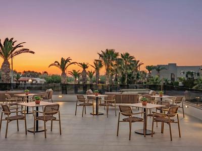 Espace extérieur vide avec tables et chaises, palmiers et coucher de soleil en arrière-plan.