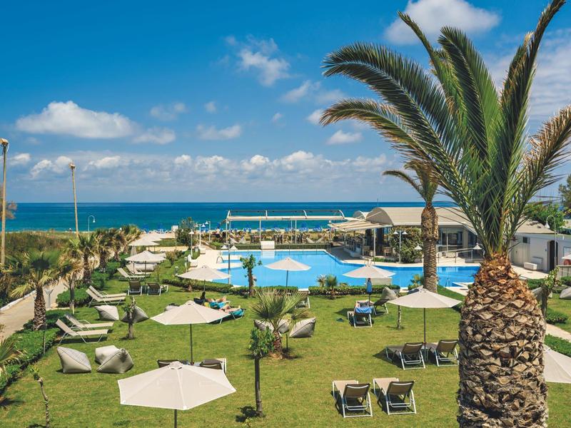 Vista de una piscina de hotel con tumbonas, sombrillas y palmeras junto al mar.