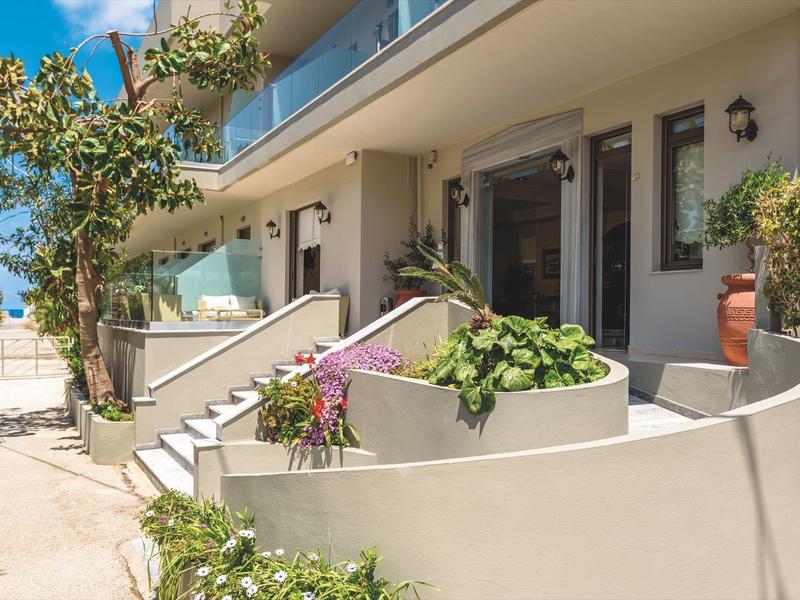 Vista exterior de un hotel moderno con balcón, escalera y plantas junto a la entrada.