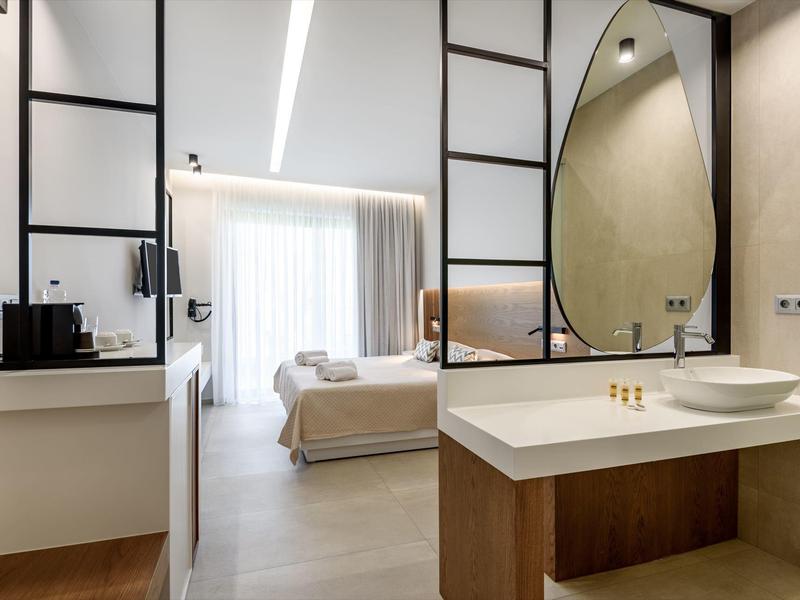 Área moderna de habitación de hotel con baño abierto, espejo redondo y vista a la cama con luz natural.