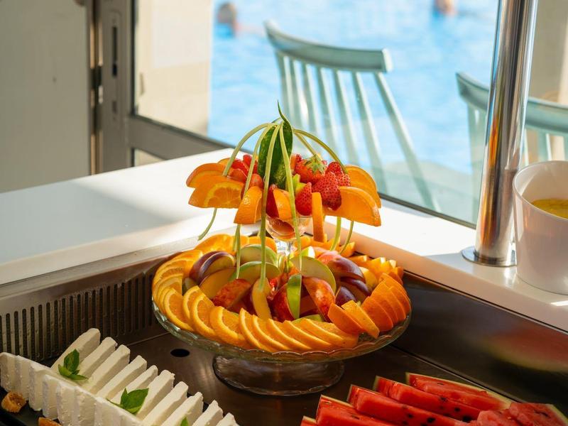 Plato colorido de frutas junto a sándwiches en un buffet con vista a una piscina.