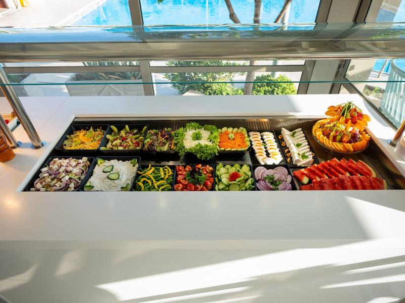 Buffet con una variedad de ensaladas coloridas y frutas junto a una piscina y palmeras.