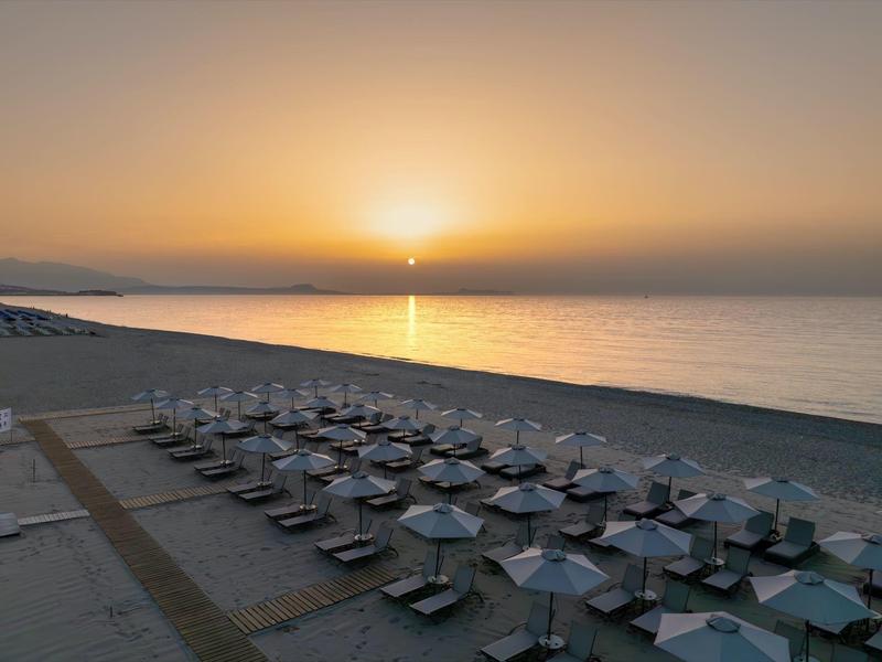 Atardecer sobre el mar tranquilo con playa vacía y filas de tumbonas y sombrillas.