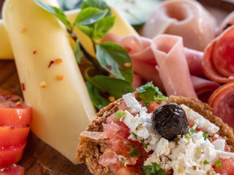 Primer plano de un plato con diferentes embutidos, quesos, tomates y rebanada de pan con queso fresco y aceituna.