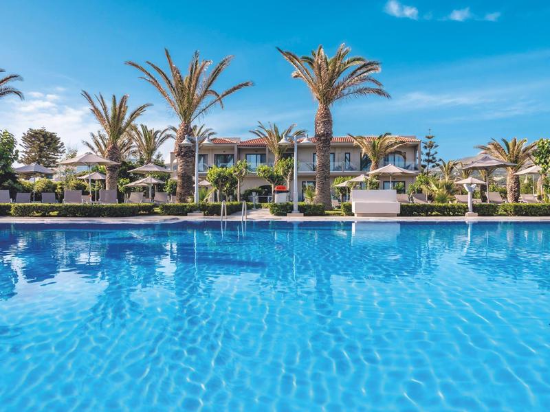 Hotel de lujo con gran piscina, palmeras y cielo azul claro al fondo.