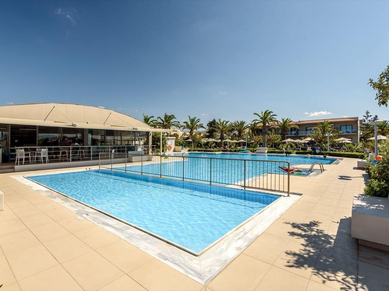 Piscina con agua clara en área exterior de hotel con palmeras y cielo azul despejado.