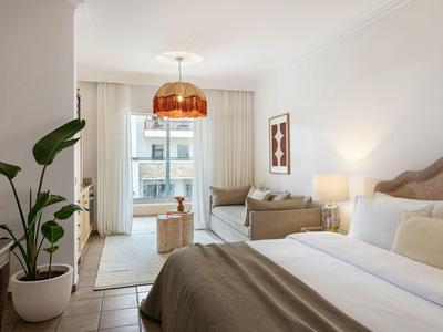 Modern eingerichteter Hotelzimmer mit Bett, Sofa, Pflanzen und großen Fenstern.