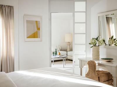 Helles Schlafzimmer mit weißer Einrichtung, Bett, Spiegel, Stuhl und dekorativen Blumen.