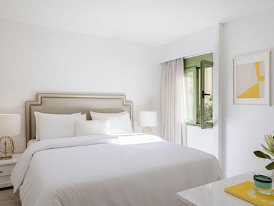 Helles Hotelzimmer mit großem Bett, weißen Wänden und offenem Fenster mit Blick ins Grüne.