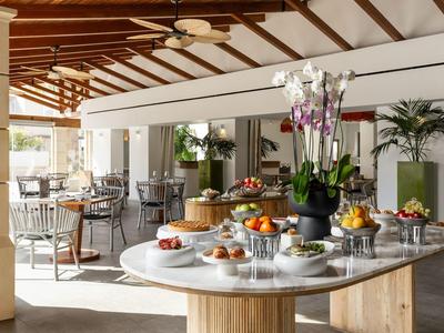 Heller Hotel-Speisesaal mit großen Tischen, Stühlen und einer Frühstücksauswahl auf dem Tisch.