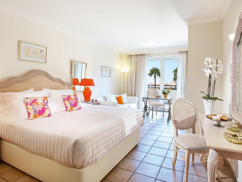 Habitación de hotel luminosa con cama grande, sofá, escritorio y balcón con vista a palmeras.