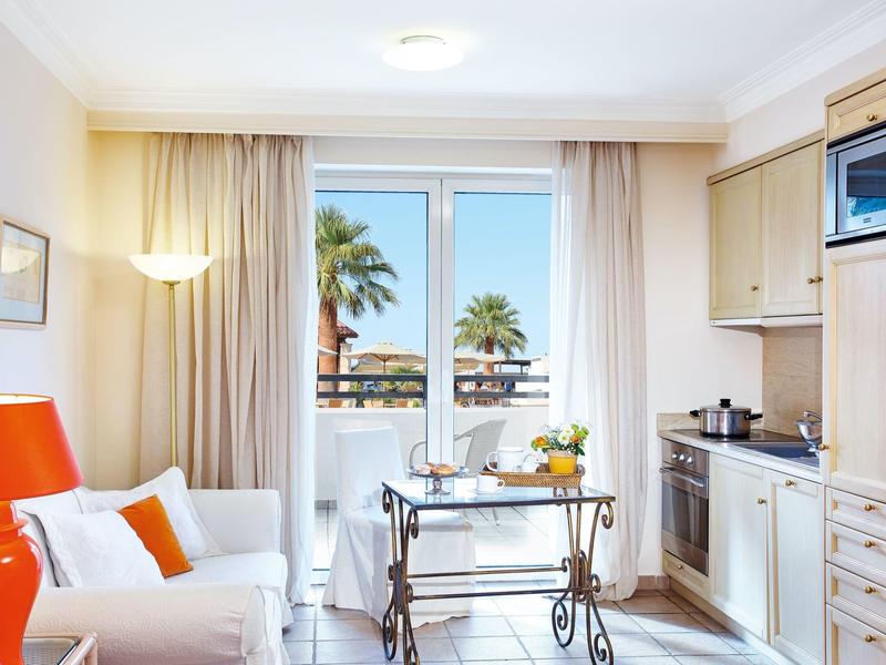 Habitación de hotel luminosa con cocina, mesa de comedor y vista a palmeras en el balcón.