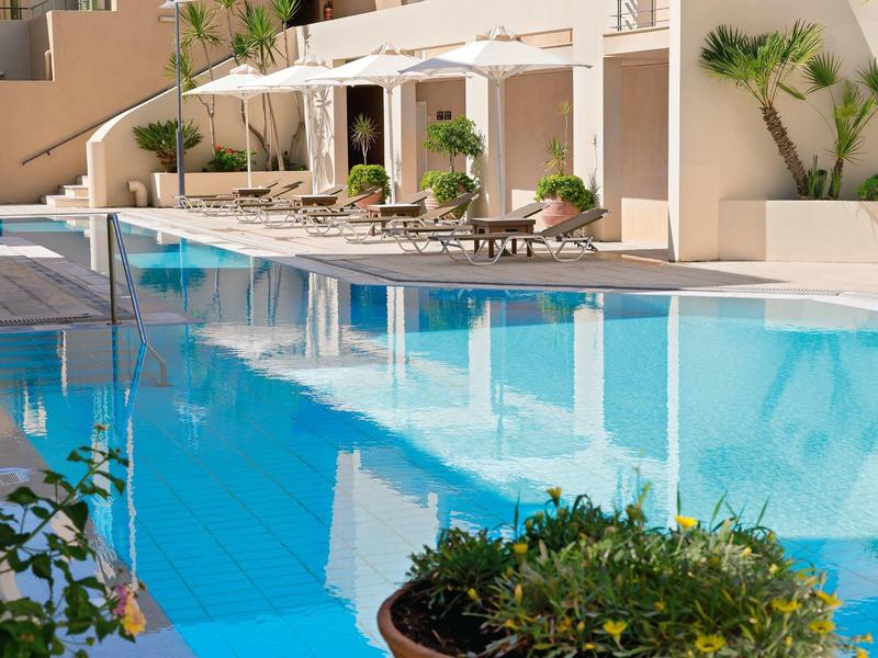 Piscina clara y grande frente a un hotel con tumbonas y palmeras, ambiente tropical y relajante.