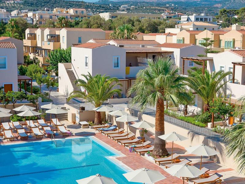 Piscina exterior con sombrillas, tumbonas y casas vacacionales en un resort tropical.