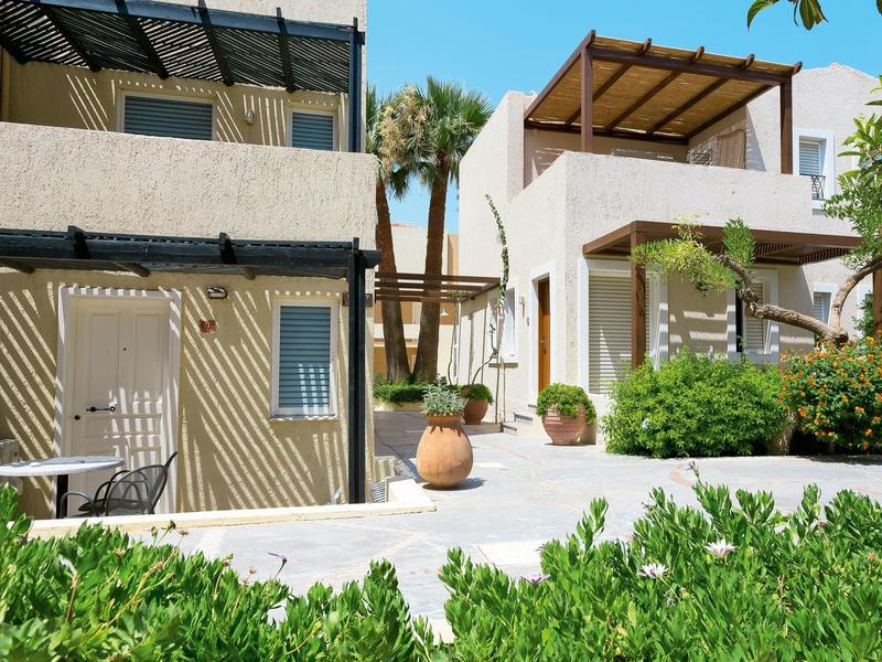 Patio de hotel moderno con área verde, toldo solar y macetas de barro mediterráneas bajo el sol.