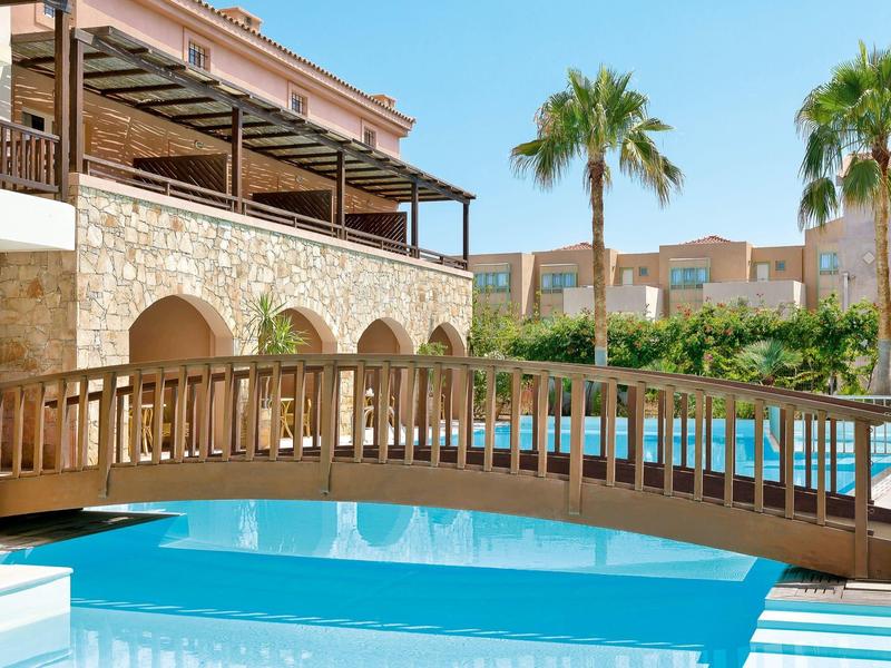 Puente de madera sobre una piscina junto a un hotel con palmeras al fondo.