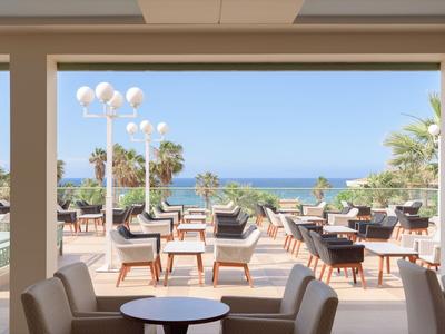 Terrasse mit Stühlen und Tischen, Blick auf Meer und Palmen unter blauem Himmel.