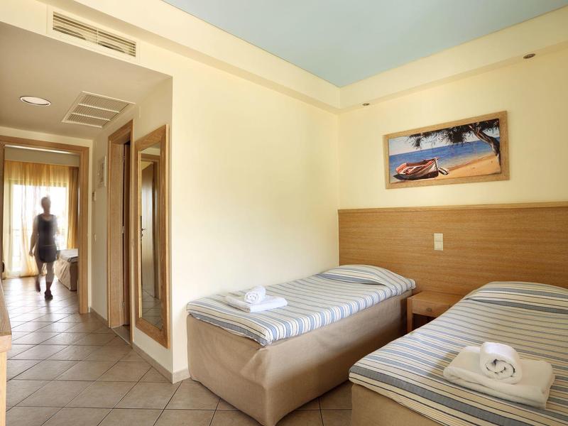 Camera d'hotel luminosa con due letti singoli e corridoio con appendiabiti.