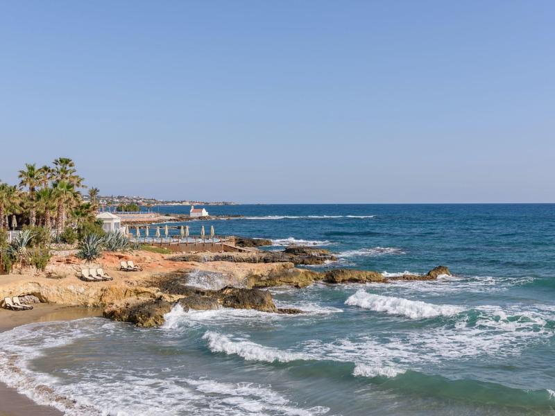 Costa rocciosa con palme, spiaggia sabbiosa e mare calmo sotto un cielo limpido.