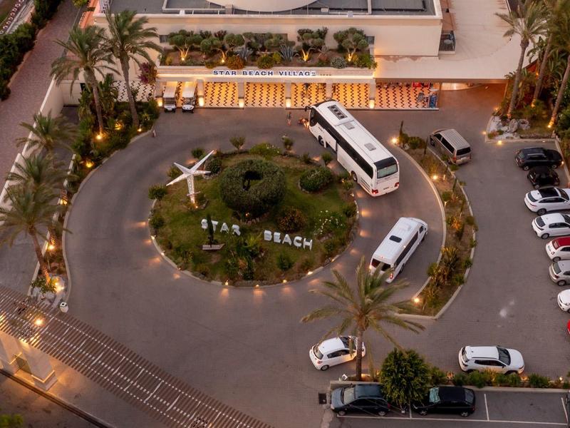 Vista aerea dell'ingresso illuminato di un hotel con rotonda, autobus e auto parcheggiate al crepuscolo.
