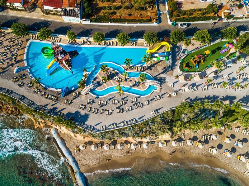 Vista aerea di un hotel con piscine, scivoli d'acqua e spiaggia con ombrelloni.