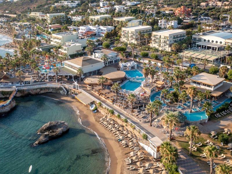 Vista aerea di un resort sulla spiaggia con piscine, palme e edifici circostanti.