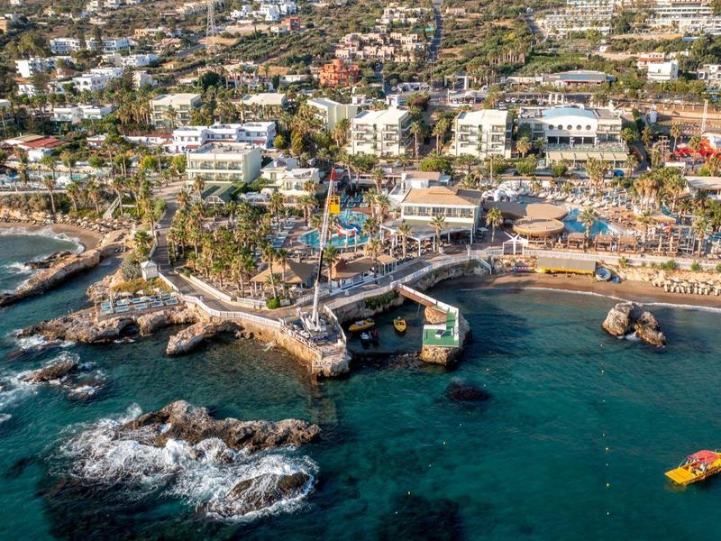 Veduta aerea di una località costiera con edifici, spiaggia, rocce e un molo nel mare.
