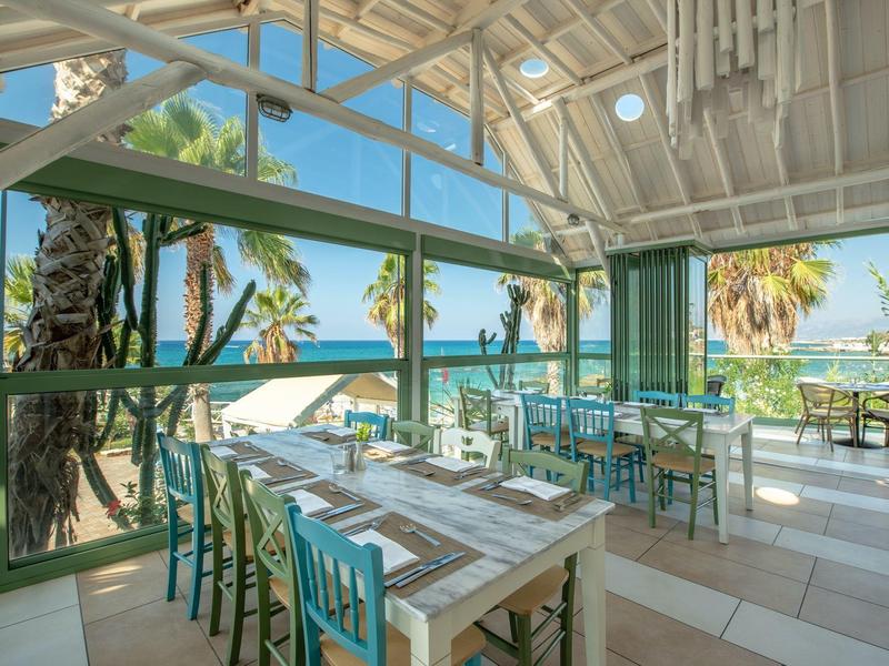 Area pranzo aperta con vista mare, tavoli in legno e sedie blu in un resort vacanze.