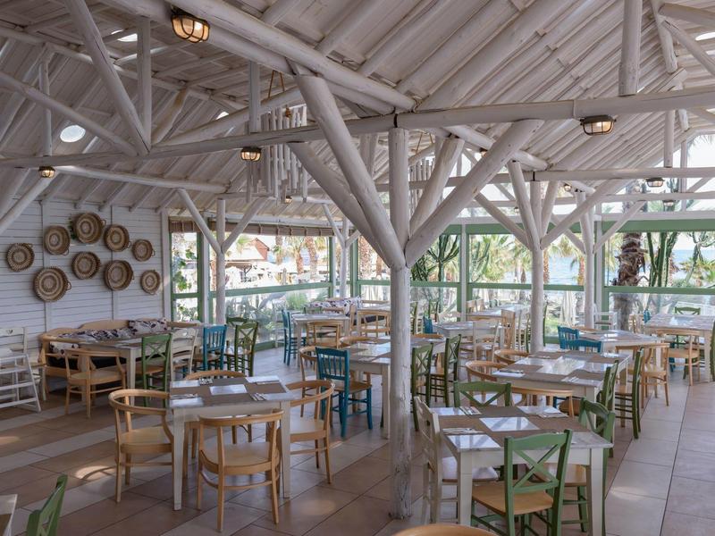 Sala da pranzo luminosa e ariosa con tavoli e sedie in legno, circondata da grandi finestre.
