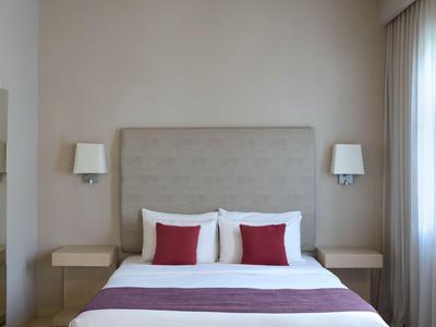 Habitación de hotel minimalista con cama doble, cabecero gris, paredes blancas y dos lámparas de pared.