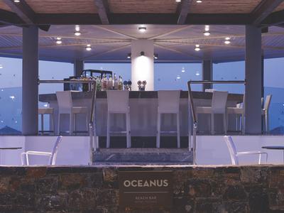 Moderní venkovní bar s barovými židličkami, skleněným zábradlím a atmosférickým osvětlením za soumraku.