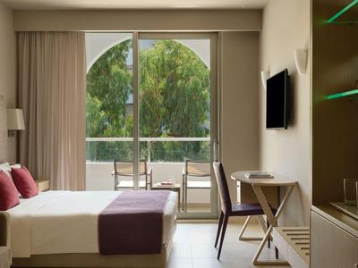 Hell eingerichtetes Hotelzimmer mit Doppelbett, Schreibtisch und Balkon mit Blick auf Bäume.