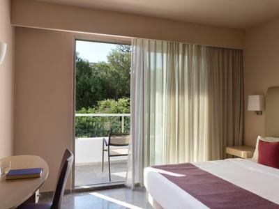 Helles Hotelzimmer mit Balkon, Vorhängen und einfacher moderner Einrichtung.