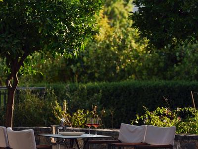 Sonnige Terrasse mit weißen Stühlen und Tischen, umgeben von grünen Bäumen und Sträuchern.