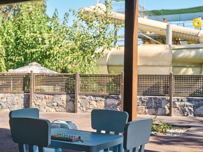 Überdachter Außenbereich mit blauem Kindertisch und Stühlen vor einer Wasserrutsche und Bäumen.