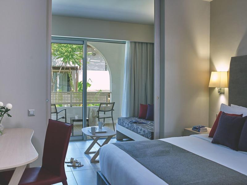 Licht ingericht hotelkamer met bed, bureau en uitzicht op balkon met tuin.