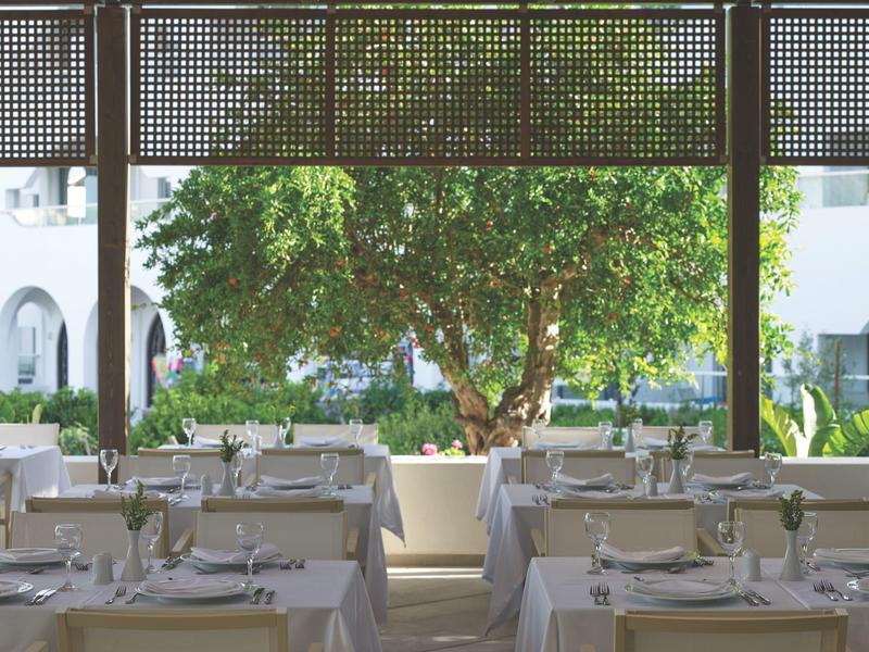 Licht restaurant met gedekte tafels en uitzicht op een boom voor een wit gebouw.