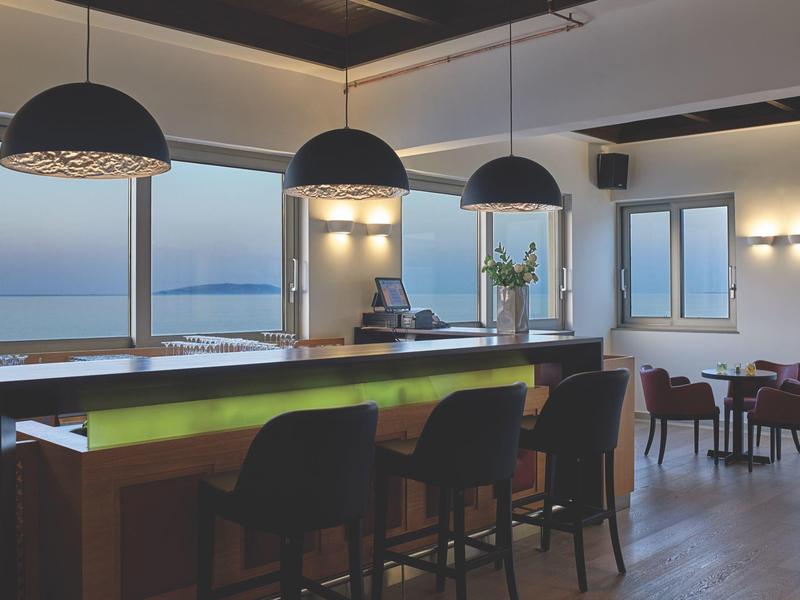 Moderne hotelbar met krukken, grote ramen en uitzicht op zee bij schemering.