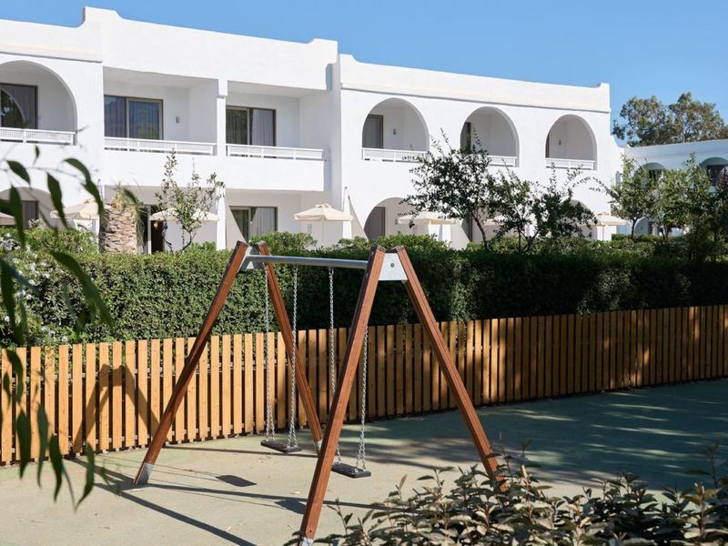 Kinderspeelplaats met schommel voor een wit hotelgebouw bij zonnig weer