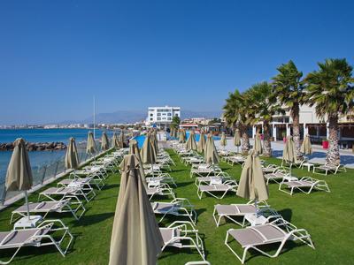 Empty sun loungers and closed umbrellas on a grassy area by the sea under clear sky.