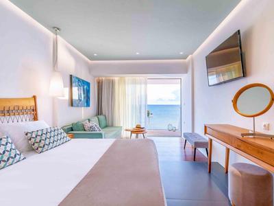 Nowoczesny pokój hotelowy z widokiem na morze, dużym łóżkiem, telewizorem i częścią wypoczynkową z sofą.