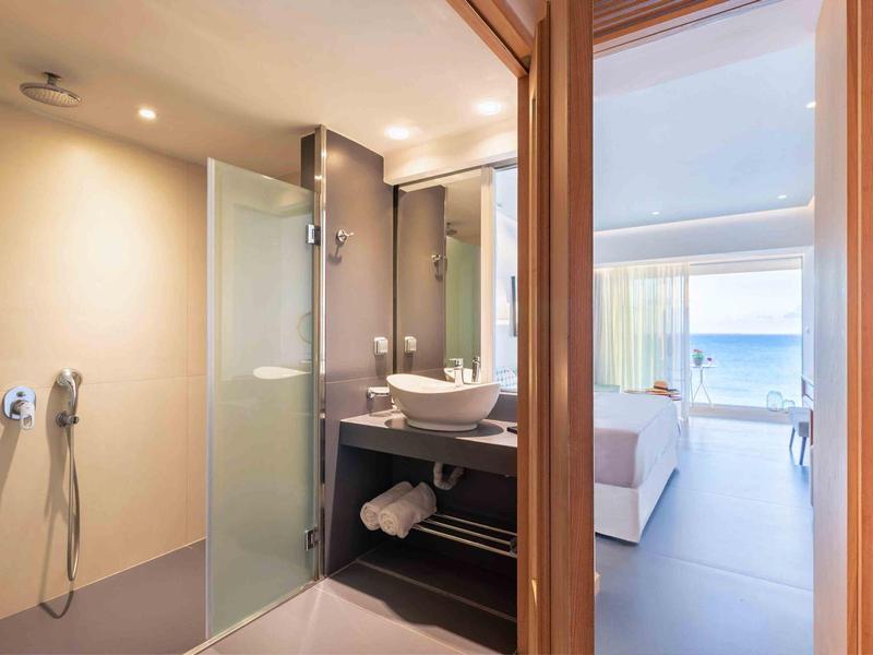 Nowoczesna łazienka hotelowa z prysznicem typu walk-in i widokiem na jasną sypialnię z widokiem na morze.