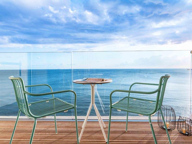 Dwa zielone krzesła i mały stolik na balkonie z widokiem na morze pod pochmurnym niebem.