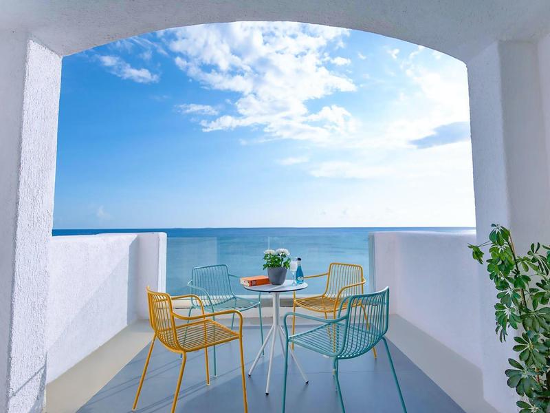 Balkon z kolorowymi krzesłami i stołem z kwiatami, widok na niebieskie morze pod niebieskim niebem.