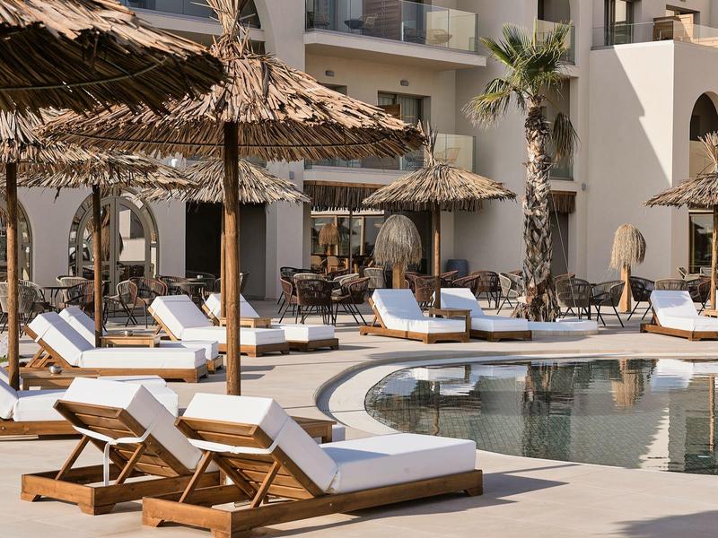 Zwembad met ligstoelen en rieten parasols bij een hotel op een zonnige dag.