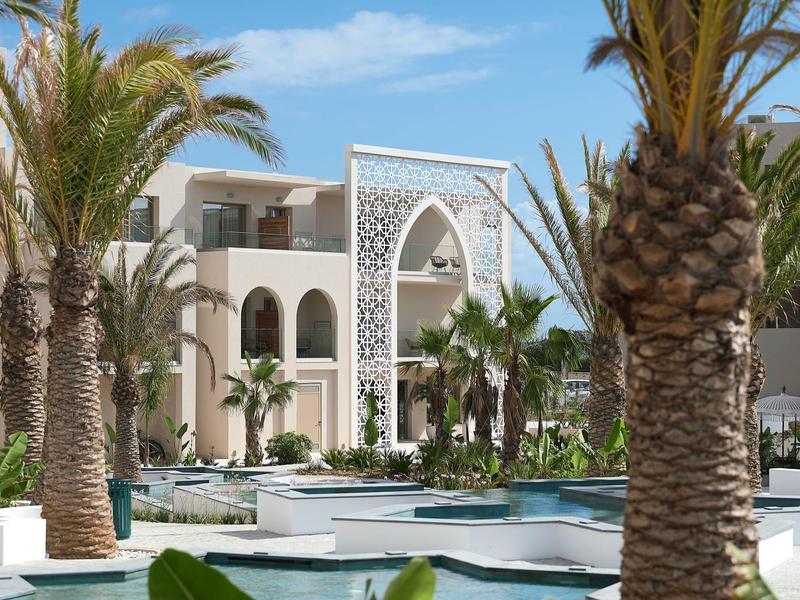 Modern hotelcomplex met zwembad en palmbomen onder een blauwe lucht.